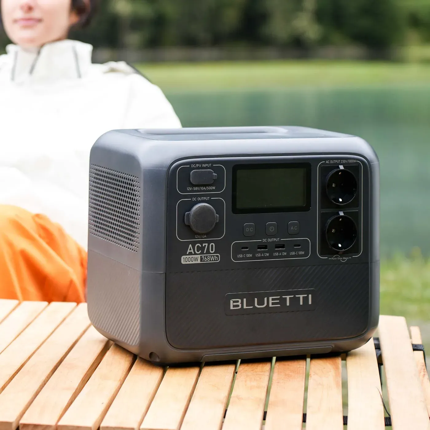 La BLUETTI AC70 posée sur une table en bois au bord d'un lac, écran allumé, une personne assise derrière