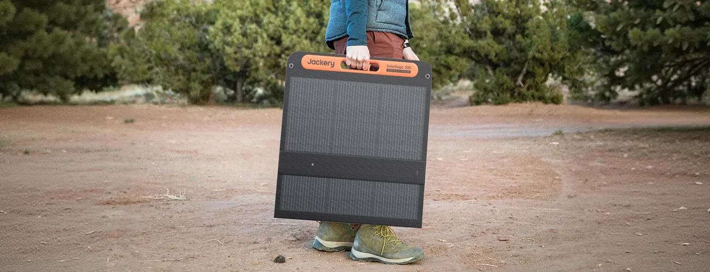 Le Jackery SolarSaga 200W plié, porté d'une main par sa poignée sur un chemin de terre